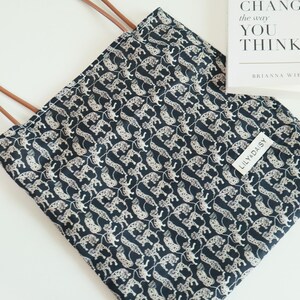 May include: A black tote bag with a white leopard print design. The bag has brown leather handles and a white label with the text "Lilly Daisy" on the front.