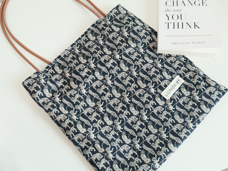 May include: A black tote bag with a white leopard print design. The bag has brown leather handles and a white label with the text "Lilly Daisy" on the front.