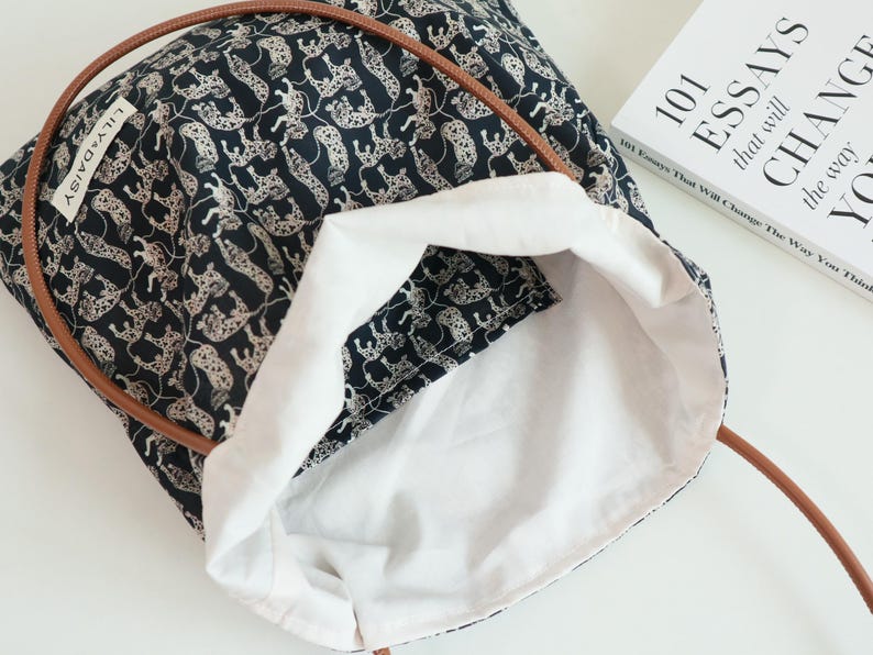 May include: A black tote bag with a white leopard print design. The bag has brown leather handles and a white cotton lining. The bag is open and the lining is visible.