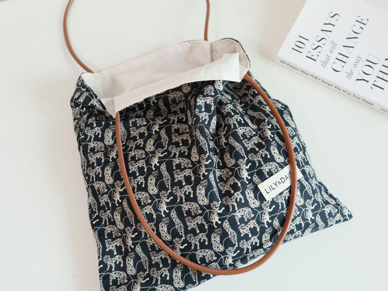May include: A black and white tote bag with a leopard print design. The bag has brown leather straps and a white cotton lining. The bag has a label that says "Lily &  Daisy".