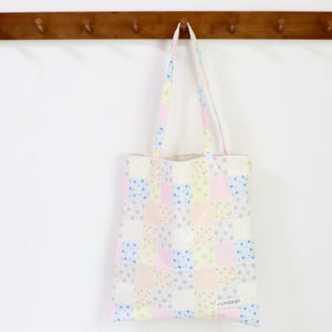 Peut inclure: Un sac fourre-tout pastel à motifs patchwork avec des anses assorties est suspendu à un porte-manteau en bois. Le sac présente un motif de carrés roses, bleus et jaunes avec de petits accents floraux. Le texte "LILANDA BY" est visible.