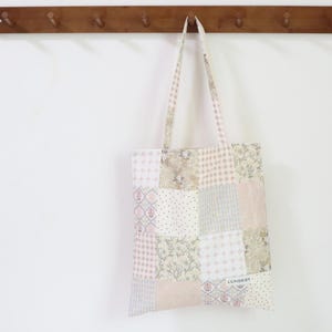 May include: A patchwork tote bag in pastel shades of pink, beige, and grey, hanging from a wooden peg rail. The bag features a mix of floral, dotted, and geometric fabric patterns.