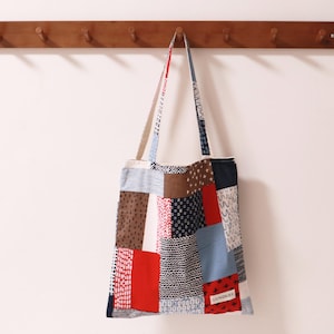 以下が含まれることがあります： 青、赤、茶色、白の様々な模様が施されたパッチワークのトートバッグ。バッグはフック付きの木製ラックに掛けられています。