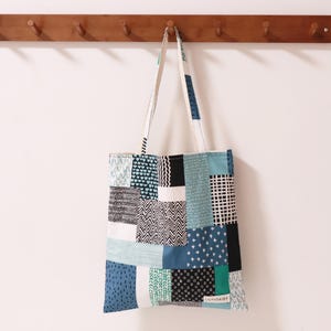 May include: A blue and white patchwork tote bag with a black and white geometric pattern. The bag is hanging on a wooden rack with hooks.