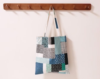 May include: A blue and white patchwork tote bag with a black and white geometric pattern. The bag is hanging on a wooden rack with hooks.
