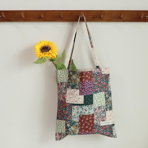 May include: A patchwork tote bag with a floral print in shades of green, pink, and brown. The bag has a brown leather handle and is hanging on a wooden rack. A sunflower is in the background.