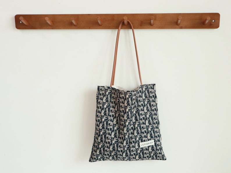 May include: A black and white tote bag with a pattern of animals hanging on a wooden wall rack with five hooks. The bag has a brown leather strap and a white label with the text "L'YNDAISY".