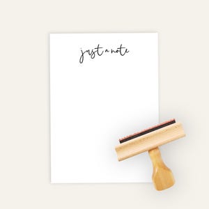May include: A white notepad with the handwritten text "just a note" in black script. A wooden stamp with a red ink pad is positioned next to the notepad, ready for use. The stamp has a light brown handle.