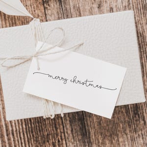 May include: A white gift box tied with twine, featuring a tag that reads "merry christmas" in elegant script. The box has a textured surface and is set against a wooden background, creating a festive holiday theme.