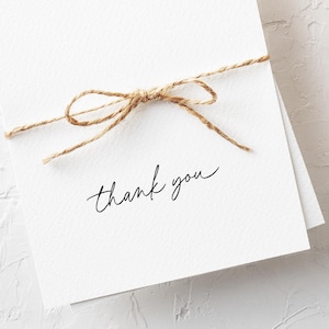 May include: A stack of white thank you cards tied with a natural twine bow. The words "thank you" are written in a delicate, cursive font on the front of the cards. The cards have a textured, matte finish.