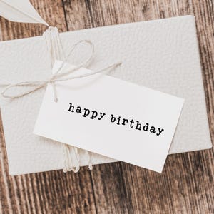 May include: A white gift box tied with twine, featuring a tag that reads "happy birthday" in black text. The box is on a wooden surface, and a white feather is tucked into the twine.