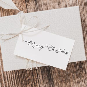 May include: A white gift box tied with twine, featuring a tag that reads "Merry Christmas" in elegant script. The box has a textured surface and rests on a wooden background, suggesting a festive holiday theme.