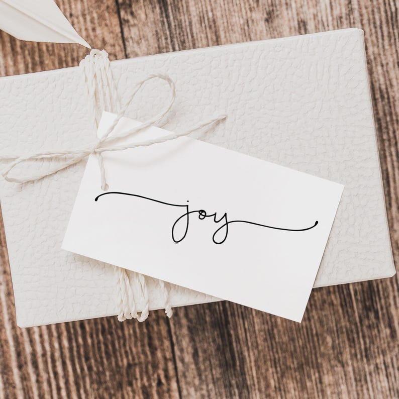 K&ouml;nnte beinhalten: Eine wei&szlig;e Geschenkbox, mit Bindfaden gebunden, mit einem wei&szlig;en Anh&auml;nger mit dem Wort "joy" in eleganter schwarzer Schrift. Eine Feder ist in den Bindfaden gesteckt. Die Box steht auf einer Holzoberfl&auml;che.