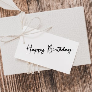 May include: A white gift box with a white tag tied with twine. The tag has the words "Happy Birthday" written in black ink.