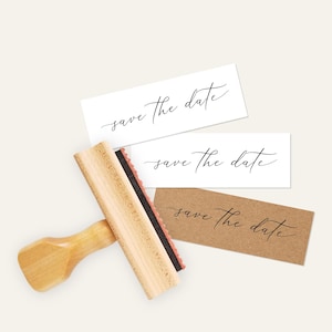May include: A wooden stamp with a black ink pad and a wooden handle. The stamp reads "save the date" in a cursive font. The stamp is shown on top of two cards, one white and one brown, with the same text printed on them.