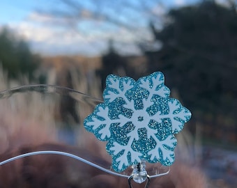 May include: A blue and white snowflake-shaped badge reel. The snowflake design has a glittery texture and is attached to a silver-colored metal clip. The background is a blurred view of a blue sky and trees.
