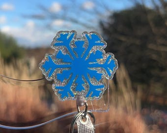 May include: A blue snowflake-shaped badge reel with silver glitter trim. The badge reel has a clear plastic base, a retractable cord, and a white lanyard with metal accents. The background shows a blue sky and out-of-focus foliage.