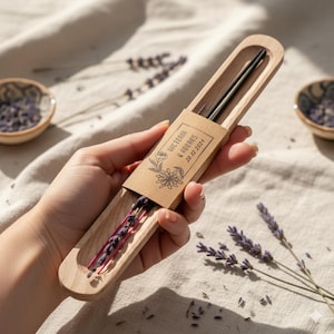 Peut inclure: Un étui en bois contenant des baguettes noires et des brins de lavande séchée. Une petite étiquette en papier brun est attachée à l'étui, avec les noms "Victoria & Andres" et la date "28.12.2021" imprimés dessus. L'étui est tenu dans une main.