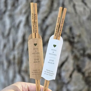 Peut inclure: Deux paires de baguettes personnalisées avec noms et date gravés, présentées dans des étuis en papier kraft et blanc. Les étuis comportent un cœur, des noms, une date et le texte "Merci d'avoir fait partie de notre histoire!"