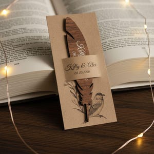 Puede incluir: Un marcapáginas de pluma de madera con los nombres "Kelly & Alex" y la fecha "08.03.2026" impresos en una tarjeta beige. El marcapáginas está colocado en un libro abierto, con una pequeña ilustración de pájaro y detalles florales.