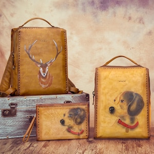 May include: A set of handcrafted leather bags and accessories. The collection includes a backpack with a deer design, a tote bag, and a wallet, each featuring a hand-painted dog portrait. The items are in shades of brown and tan.