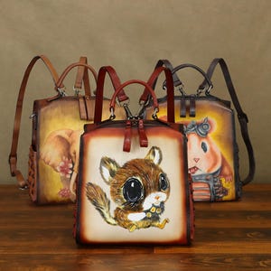 Può includere: Tre zaini in pelle con illustrazioni di animali. Lo zaino anteriore presenta uno scoiattolo con grandi occhi. Gli altri due zaini hanno un design di elefante e criceto. Ogni borsa ha spallacci regolabili e una maniglia superiore.