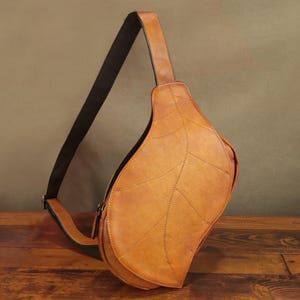 Könnte beinhalten: Eine hellbraune Leder-Umhängetasche in Blattform mit schwarzem Riemen. Die Tasche hat aufgenähte Details, die Blattadern nachahmen. Die Tasche befindet sich vor einem braunen Hintergrund und liegt auf einer Holzoberfläche.