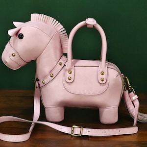 May include: A light pink horse-shaped handbag with a top handle and a detachable shoulder strap. The bag features a zipper closure, gold-colored hardware, and a tassel. The horse design includes a mane, ears, and a detailed face.