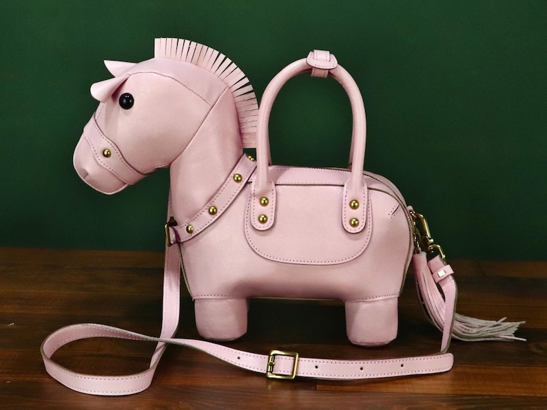May include: A light pink horse-shaped handbag with a top handle and a detachable shoulder strap. The bag features a zipper closure, gold-colored hardware, and a tassel. The horse design includes a mane, ears, and a detailed face.