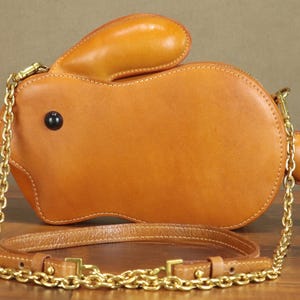 May include: A tan leather crossbody bag shaped like a rabbit. The bag features a long gold chain strap and a shorter leather strap. The rabbit has black button eyes and a small round tail. The bag is sitting on a wooden surface.