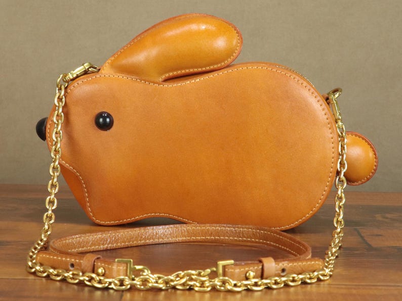 May include: A tan leather crossbody bag shaped like a rabbit. The bag features a long gold chain strap and a shorter leather strap. The rabbit has black button eyes and a small round tail. The bag is sitting on a wooden surface.