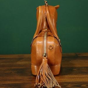 May include: A tan leather handbag shaped like a horse, viewed from the rear. The bag features a zipper closure, a tassel tail, and small details like a horse's ears and a mane. The bag is on a wooden surface.