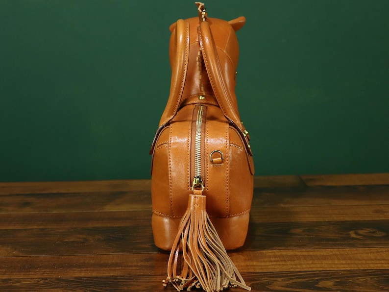 May include: A tan leather handbag shaped like a horse, viewed from the rear. The bag features a zipper closure, a tassel tail, and small details like a horse's ears and a mane. The bag is on a wooden surface.