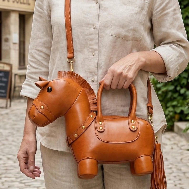May include: A brown leather handbag shaped like a horse, featuring a detailed head, mane, and legs. The bag has a top handle, a shoulder strap, and a decorative tassel. Gold-colored hardware accents the design.