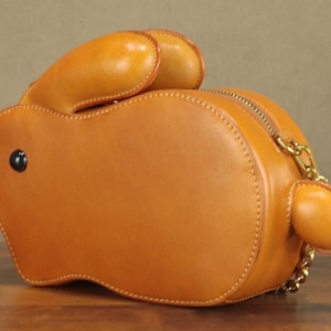 May include: A tan leather handbag shaped like a rabbit. The bag features a zipper closure, a gold chain strap, and a black button eye. The rabbit's ears and tail are also made of tan leather. The bag is approximately 10 inches long.