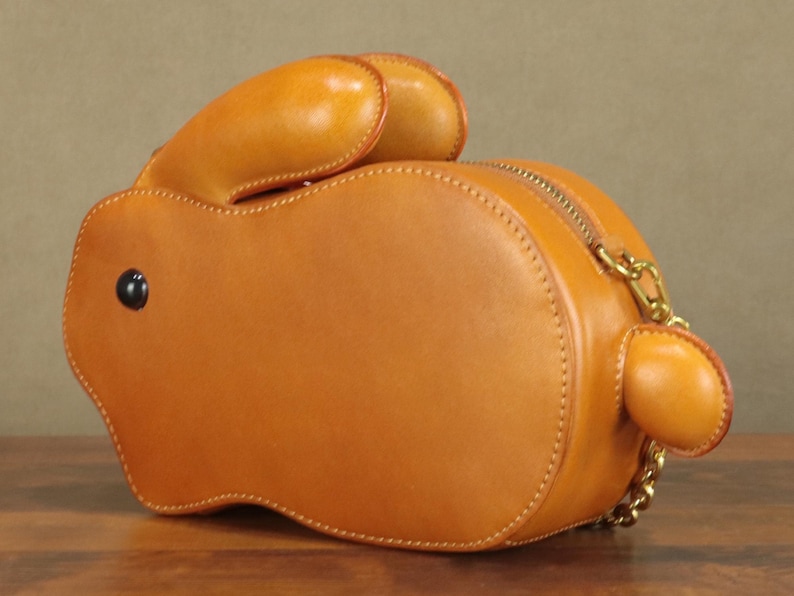 May include: A tan leather handbag shaped like a rabbit. The bag features a zipper closure, a gold chain strap, and a black button eye. The rabbit's ears and tail are also made of tan leather. The bag is approximately 10 inches long.