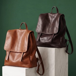May include: Two leather backpacks, one in a light tan color and one in a dark brown color. Each backpack features a large fold-over flap, a top handle, and adjustable shoulder straps. The bags have a smooth finish and vertical seam detailing.