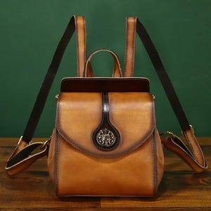 May include: A small, brown leather backpack with adjustable straps. The bag features a flap closure with a decorative metal emblem and a top handle. The leather has a distressed appearance, and the straps are black.