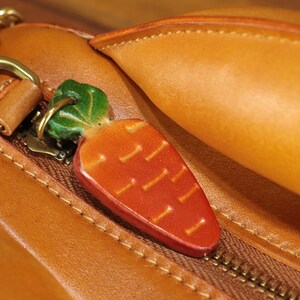 May include: A close-up of a leather bag with a carrot-shaped charm. The charm is orange with green leaves and is attached to the zipper. The bag is brown with a gold zipper and hardware.