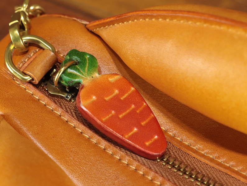 May include: A close-up of a leather bag with a carrot-shaped charm. The charm is orange with green leaves and is attached to the zipper. The bag is brown with a gold zipper and hardware.