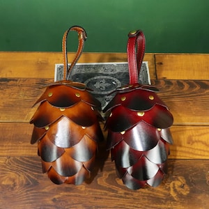 May include: Two leather pinecone-shaped pouches with handles. One is a gradient of brown, the other is red. Both have brass rivets and a leather loop handle. The pouches are on a wooden surface.