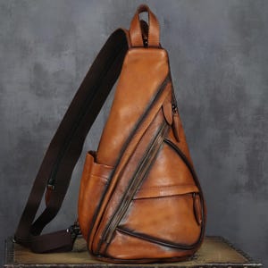Puede incluir: Bolso bandolera de cuero marrón con una sola correa ajustable para el hombro. El bolso cuenta con un compartimento principal con cremallera, un bolsillo exterior y un asa superior. El cuero tiene un aspecto envejecido con bordes más oscuros y herrajes marrones oscuros.