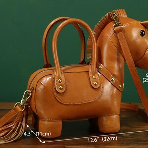 May include: A brown leather handbag shaped like a horse, with a zipper closure and two top handles. The bag has a detachable shoulder strap and a tassel. The bag is 12.6 inches (32 cm) long, 4.3 inches (11 cm) wide, and 9.8 inches (25 cm) tall.