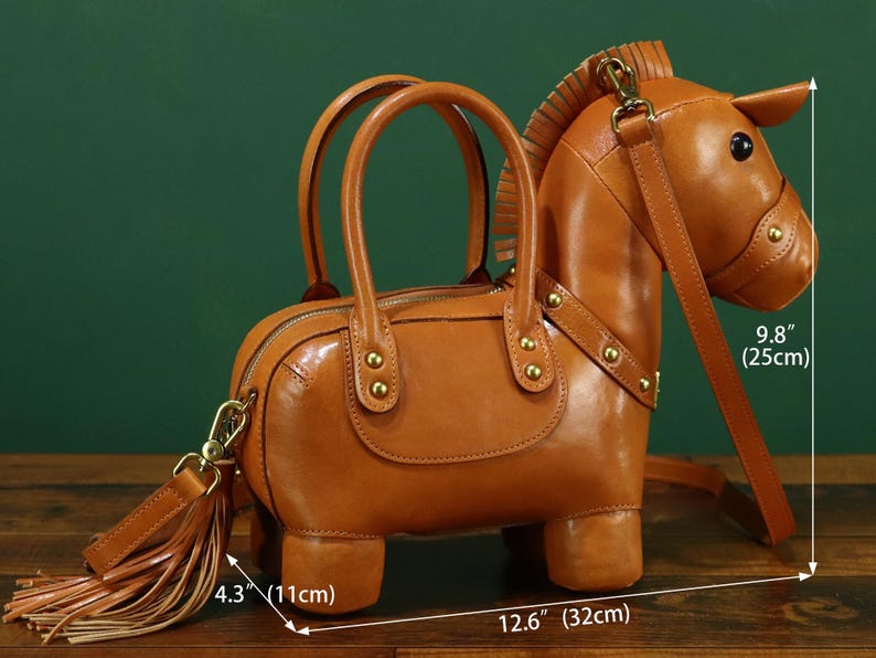 May include: A brown leather handbag shaped like a horse, with a zipper closure and two top handles. The bag has a detachable shoulder strap and a tassel. The bag is 12.6 inches (32 cm) long, 4.3 inches (11 cm) wide, and 9.8 inches (25 cm) tall.