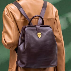May include: A dark brown leather backpack with a structured frame, a top handle, and adjustable shoulder straps. The bag features a gold-toned metal push-lock closure on a central vertical strap, a textured surface, and a wide, rectangular opening.