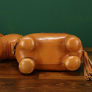May include: A brown leather handbag shaped like a hippopotamus, with four rounded feet and a tassel. The bag is lying on a wooden surface with a green background. The bag has a zipper closure and a small handle.