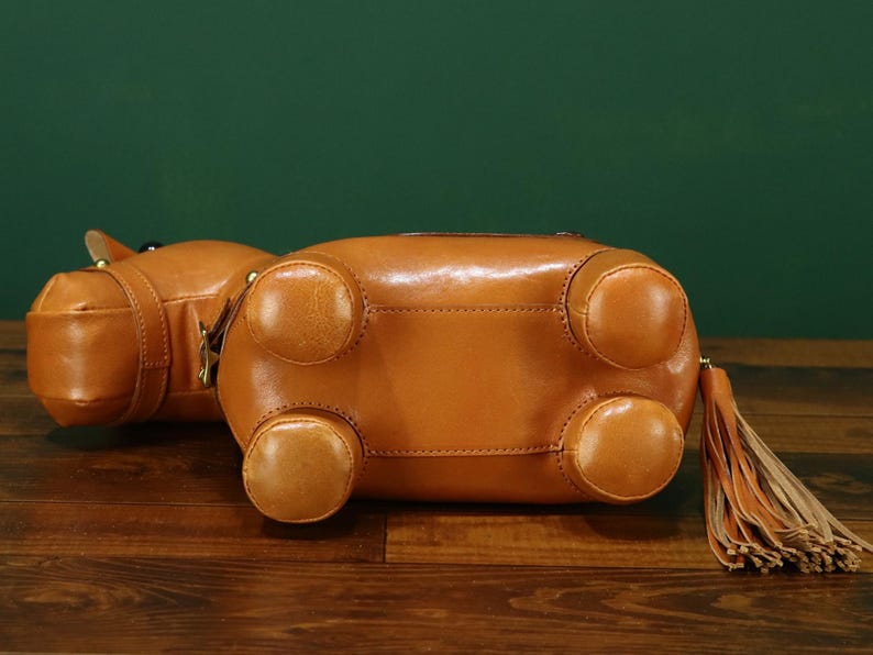 May include: A brown leather handbag shaped like a hippopotamus, with four rounded feet and a tassel. The bag is lying on a wooden surface with a green background. The bag has a zipper closure and a small handle.