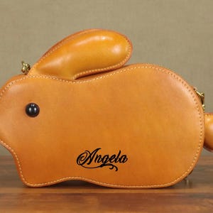 May include: A tan leather bunny-shaped handbag with a zipper closure and a small handle. The bag features a black eye and the name "Angela" in cursive script. The bag is made of stitched leather.