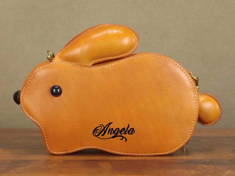 May include: A tan leather bunny-shaped handbag with a zipper closure and a small handle. The bag features a black eye and the name "Angela" in cursive script. The bag is made of stitched leather.