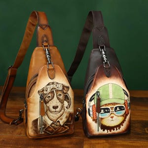 May include: Two leather sling bags. One is tan with a dog wearing aviator goggles and a jacket. The other is brown with a cat wearing a green cap, sunglasses, and headphones. Both bags have a zip and adjustable strap.